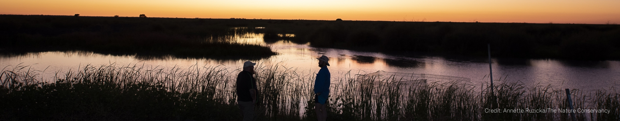 Two men stand silhouetted against a brilliant purple and orange sunset overlooking water on the conservation area of Gayini Nimmie Caira 