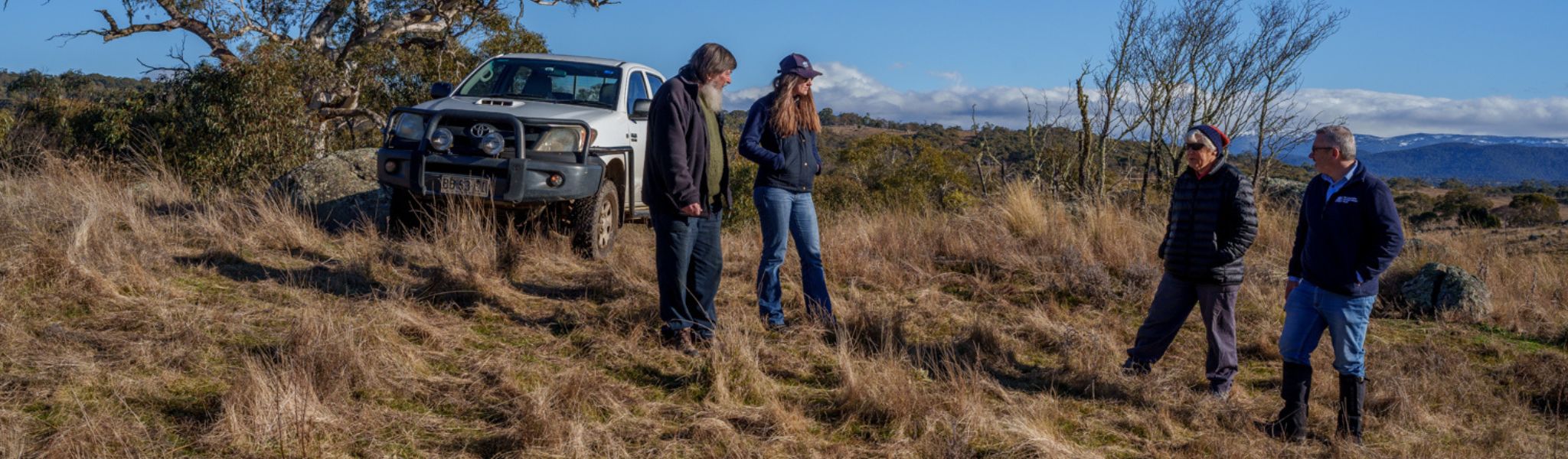 Four people stand aside a vehicle in a grassy and wooded paddock of a rural property