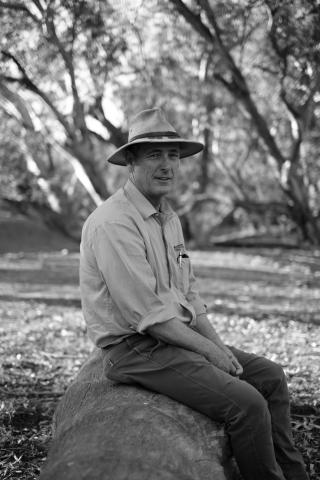 Black and white picture of a white man with a hat sitting on a rock in the bush 